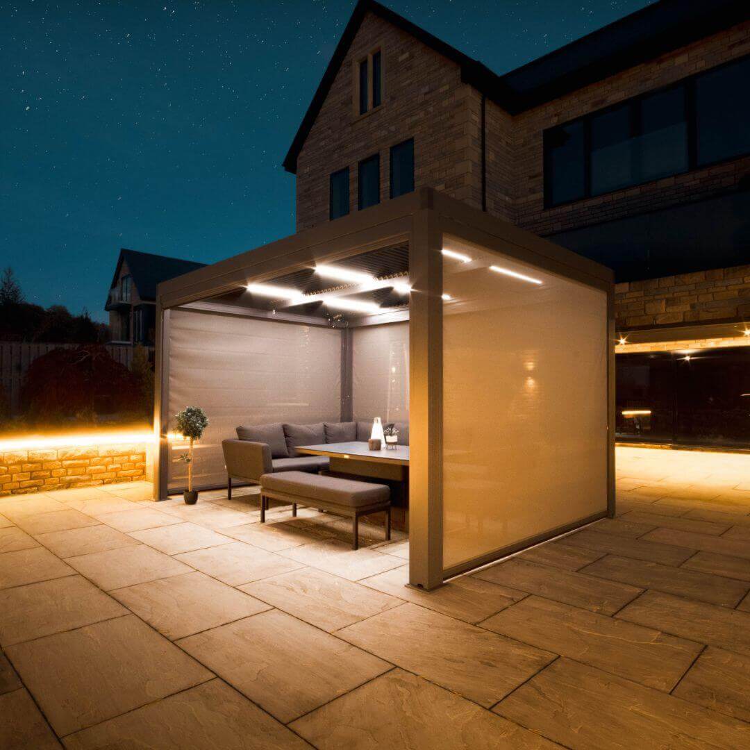 Side view of a grey aluminium pergola with louvred roof. The white LED lights underneath the roof panel are turned on. Three Textilene privacy screens attached to the pergola are pulled down.
