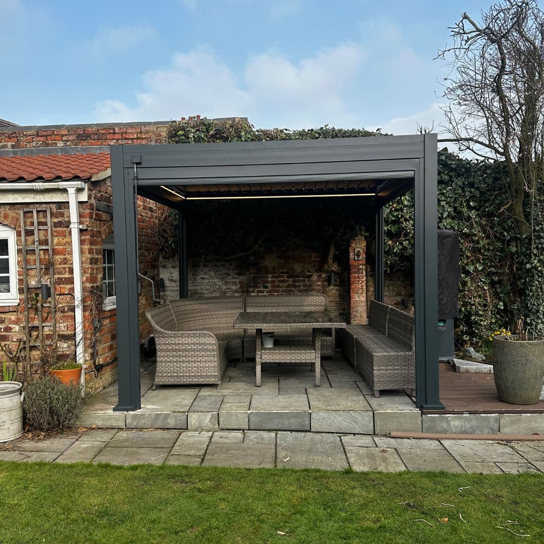 Front view of a grey aluminium pergola with louvred roof. The white LED lights underneath the roof panel are turned on.