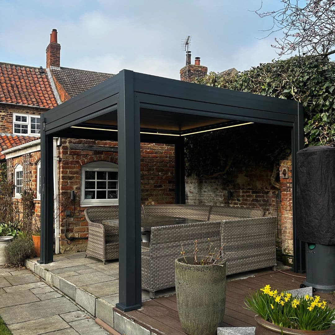 Front view of a grey aluminium pergola with louvred roof. The white LED lights underneath the roof panel are turned on.