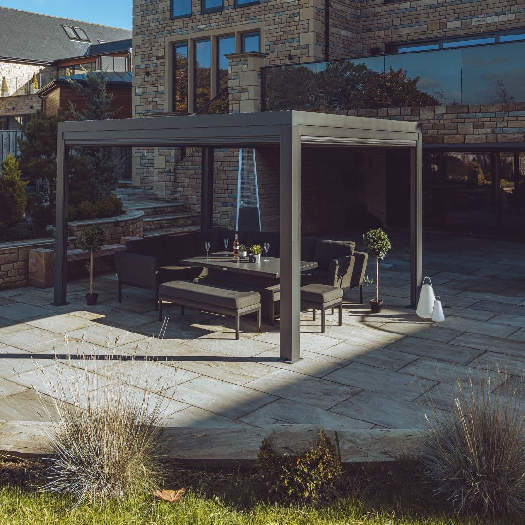 Side view of a grey aluminium pergola with louvred roof. An outdoor fabric corner dining set sits under the pergola.