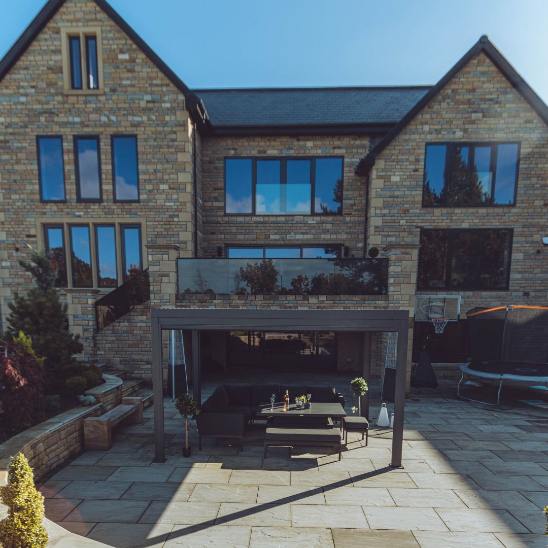 Side view of a grey aluminium pergola with louvred roof. An outdoor fabric corner dining set sits under the pergola.