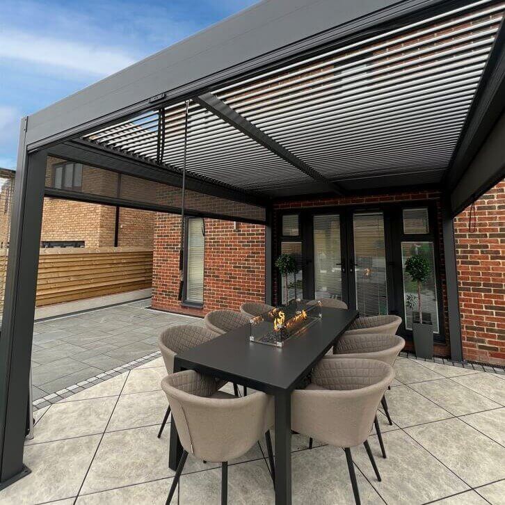 Close up of an anthracite grey aluminium pergola with manual louvred roof. A fire pit rectangle dining set sits under the pergola.