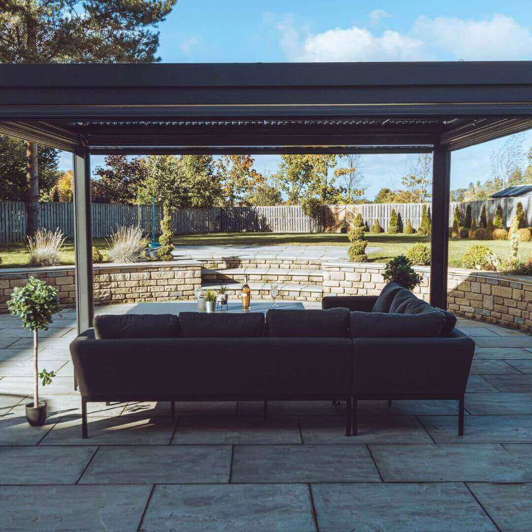 Close up of a grey aluminium pergola with louvred roof frame. An outdoor fabric corner dining set sits under the pergola.