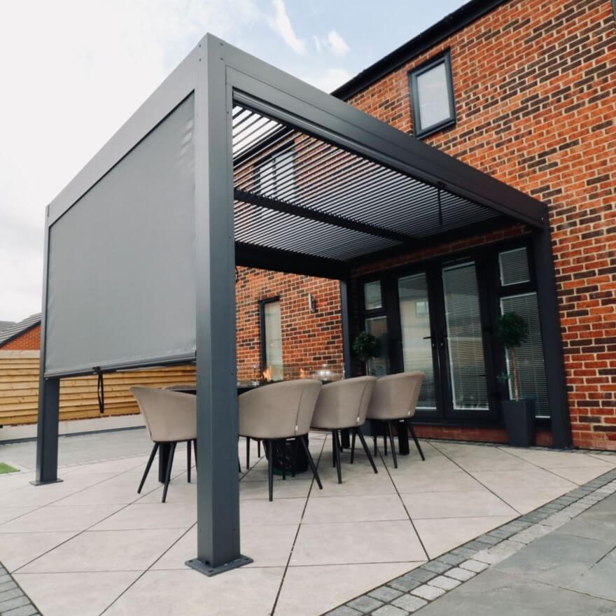 Side view of a grey aluminium pergola with louvred roof. A Textilene privacy screen is attached to the pergola and pulled down.