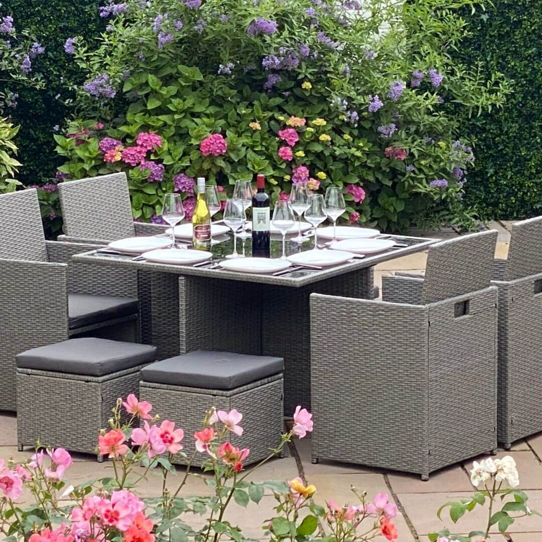 Light grey rattan cube dining set with a square dining table, 4 dining chairs and 4 stools. Tableware and bottles of wine are on top of the table.