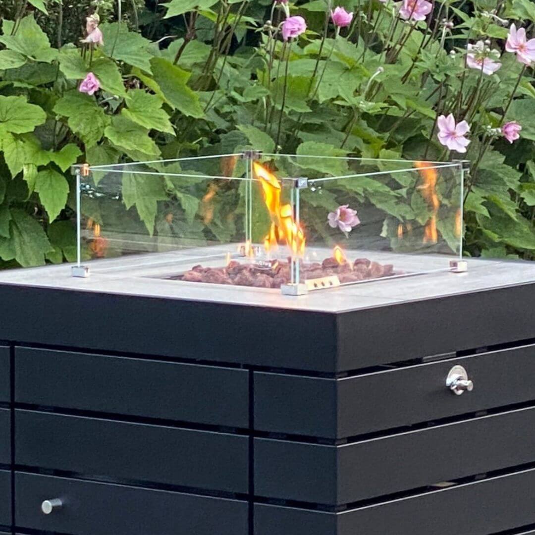 Close up of a dark grey aluminium square fire pit with wind guard. The fire pit is turned on.