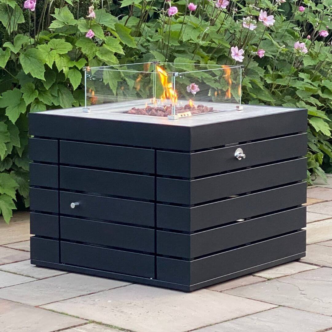 A dark grey aluminium square fire pit with wind guard. The fire pit is turned on.