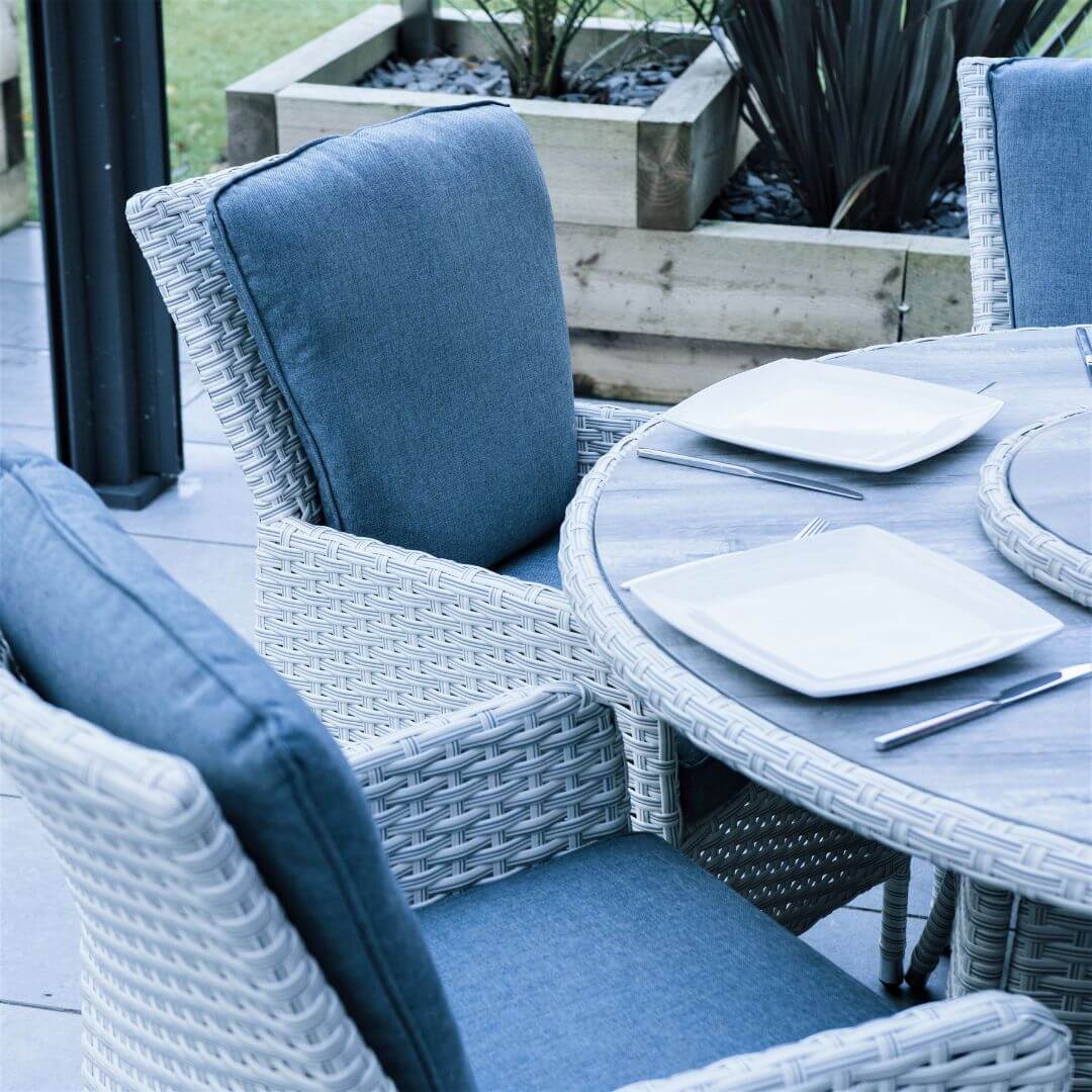 Close up of two light grey rattan dining chairs with padded back and seat cushions and a round dining table with ceramic printed glass table top.