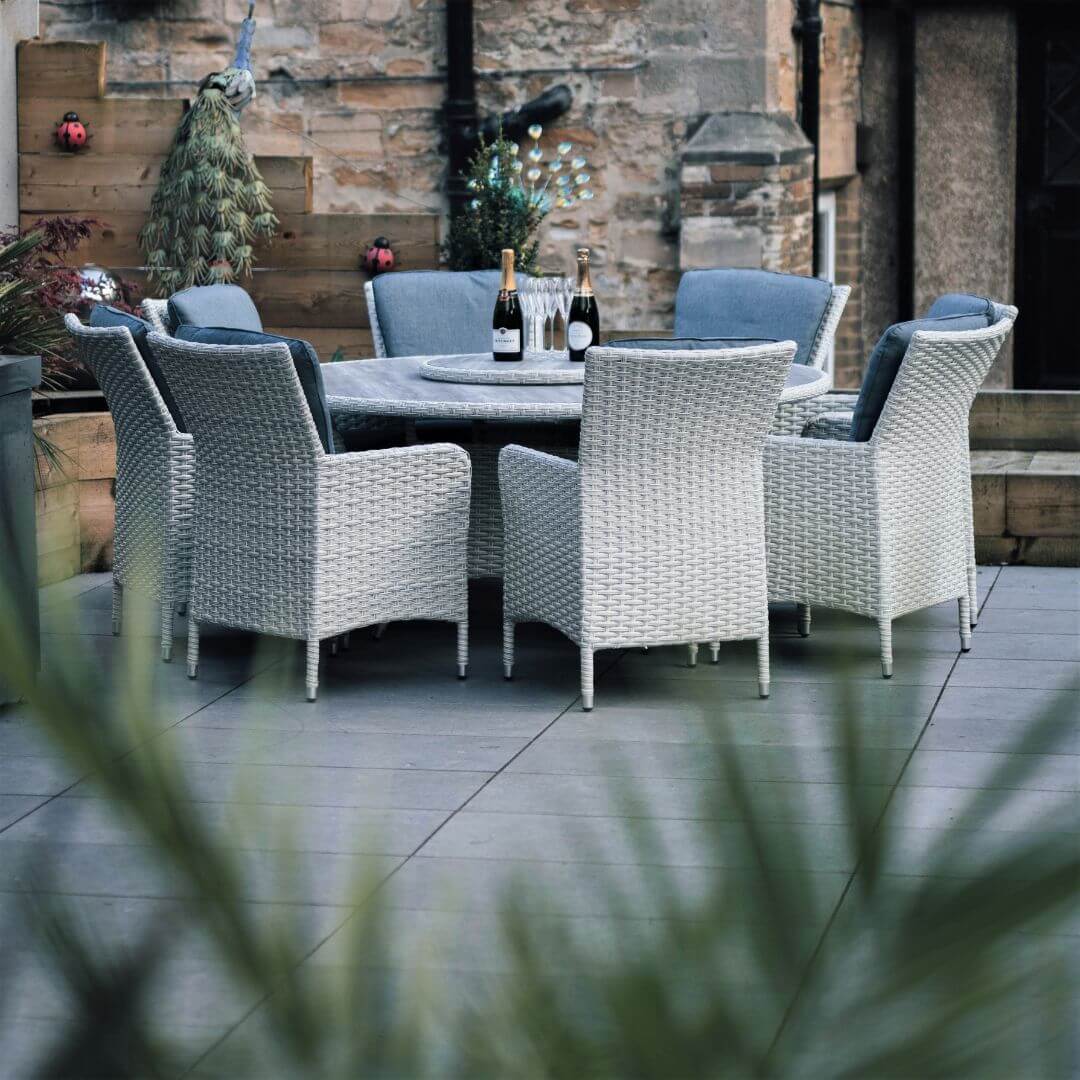 Side view of a light grey rattan 8 seat round dining set with ceramic printed glass table top and lazy susan. Green plants in the foreground.