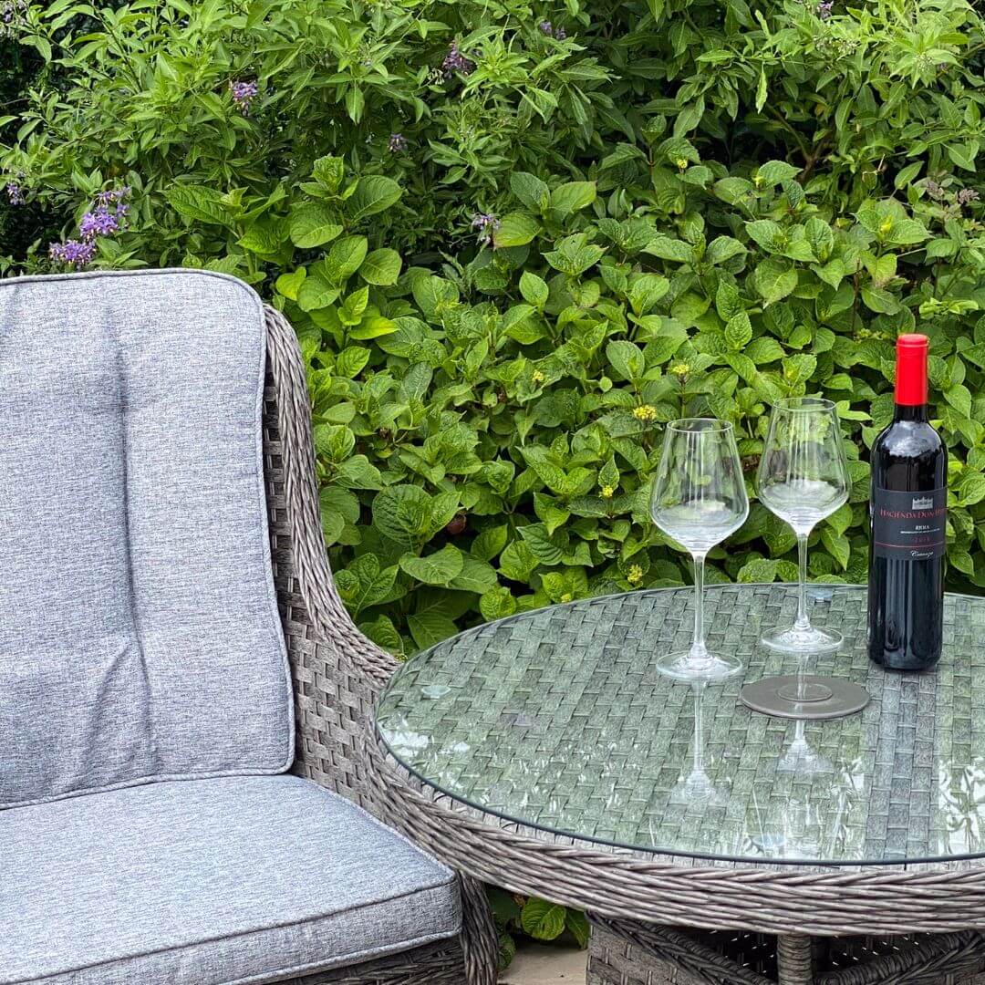 Close up of a dark grey rattan armchair and a small round table with tempered glass table top. A bottle of wine and two glasses on top of the table.