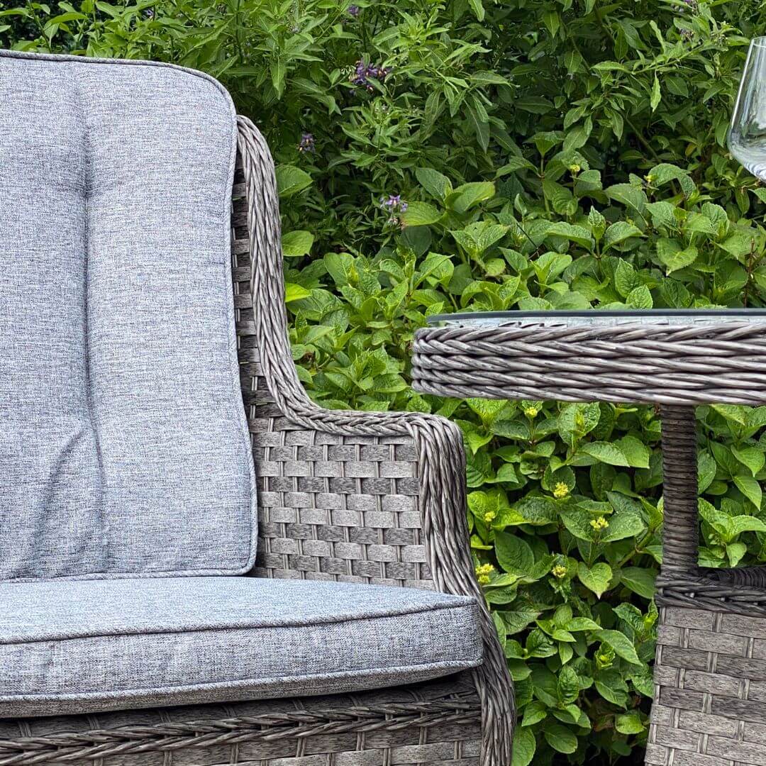 Close up of a dark grey rattan armchair with padded back and seat cushions. Side view of a small round table with tempered glass table top.