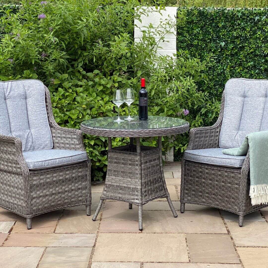 Dark grey rattan 2 seat bistro set with two armchairs and a small round table. Padded back and seat cushions on the armchairs. A bottle of wine and two glass on top of the table.